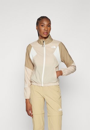 Giacca leggera beige con accenti bianchi, colletto alto, chiusura con zip e polsini elastici; abbinata a pantaloni beige, design minimalista.