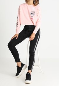 Camisa corta de manga larga rosa con varios logotipos impresos y jeans ajustados negros con rayas laterales blancas. Combinado con deportivas negras.