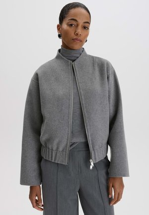 Donna che indossa una giacca grigia con zip sopra un dolcevita grigio e pantaloni grigi, in piedi davanti a uno sfondo chiaro e uniforme.