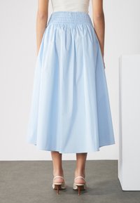 Jupe mi-longue bleu clair avec une taille smockée froncée. Tissu lisse et léger. Associée à des sandales à talons avec des lanières.