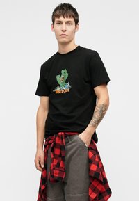 Melna kokvilnas T-kreklu, kurā ir zaļš attēls ar roku un krāsainiem elementi, ar "SANTA CRUZ" oranžā krāsā. Pelēki bikses ar sarkanu un melnu šķērsotā raksta kreklu, kas sasiets ap vidukli.