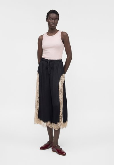 Γυναίκα φοράει ένα ανοιχτό ροζ αμάνικο τοπ, μαύρο παντελόνι με φαρδιές παντελόνες και μπεζ δαντελένιες λεπτομέρειες, και κόκκινα παπούτσια Mary Jane, στέκεται μπροστά σε λευκό φόντο.