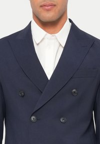 Σταυρωτό σακάκι ναυτικού μπλε με ομαλή υφή, με δύο μεγάλα μαύρα κουμπιά και γιακά με χαραγή, φορεμένο πάνω από λευκό πουκάμισο.