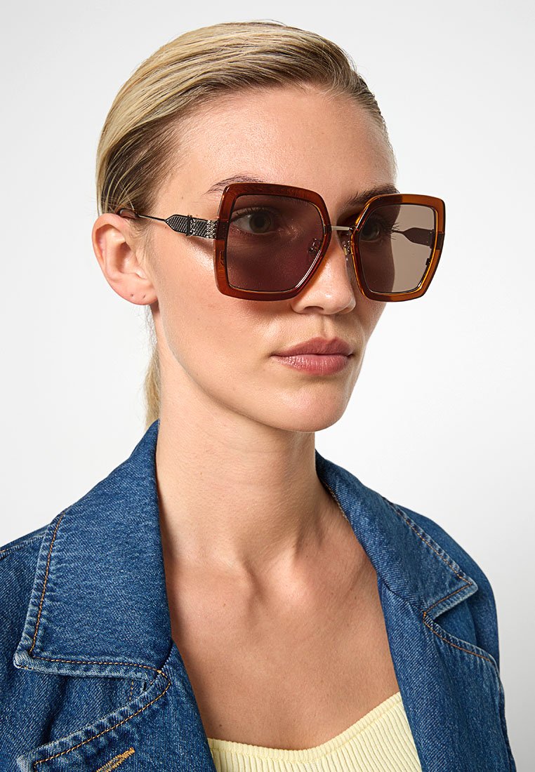 Lunettes de soleil carrées marron surdimensionnées avec des verres teintés et un accent métallique sur les branches, portées par une personne en veste en denim.