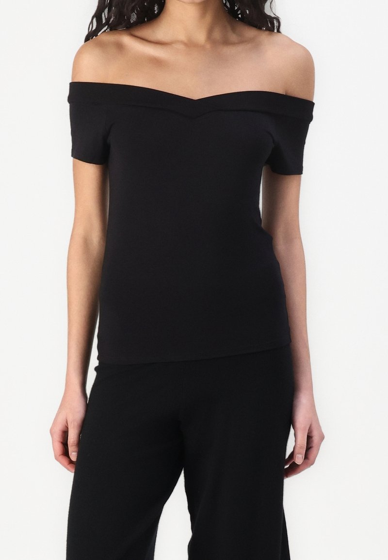 Femme portant un haut noir ajusté épaules dénudées et un pantalon noir, vue du cou aux hanches sur fond blanc.