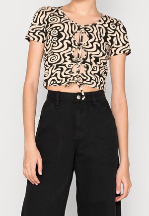 Mujer con un crop top de patrón abstracto beige y negro con aberturas delanteras y detalle de lazo, combinado con pantalones negros de talle alto.