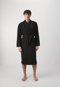 Robe de bain noire en éponge avec un design enveloppant, une ceinture et deux poches latérales. Présente le logo rouge "HUGO" sur la poitrine gauche. Texture douce.
