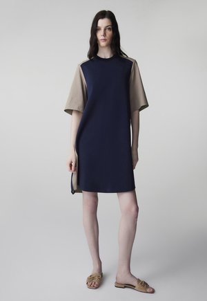 Frau steht und trägt ein marineblau-beiges Kleid mit kurzen Ärmeln und seitlichen Schlitzen sowie beigefarbene Slip-On-Sandalen, vor einem schlichten grauen Hintergrund.