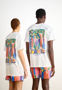 Witte T-shirts met kleurrijke, gedetailleerde prints van loofbomen en pilaren op de achterkant, gecombineerd met gestreepte en bloemige shorts.