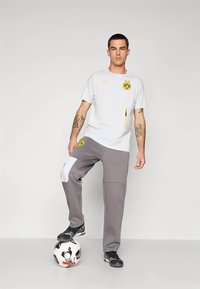 Camisa desportiva branca da Puma com logotipo preto e amarelo, combinada com calças de treino cinzentas com um remendo branco e bolso com fecho. Ténis pretos.