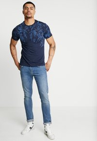Navy blue t-shirt with a textured leaf pattern, paired with light blue denim jeans and white sneakers with black accents.