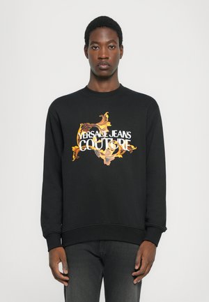 Svart hettegenser laget av bomull, med en iøynefallende, fargerik grafikk av et blomstermotiv og teksten "VERSACE JEANS COUTURE" i hvitt.