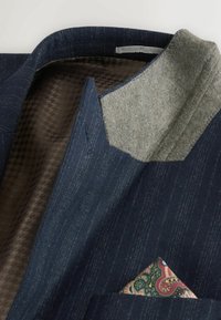 Sötétkék blézer finom csíkozással, gyapjú gallérral és mintás zsebkendővel. Belseje texturált barna béléssel rendelkezik, ami kutyafog mintázatú.