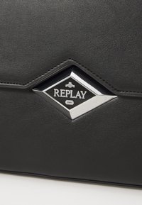 Zwarte neplederen tas met een diamantvormig zilver en zwart logo met de inscripties "REPLAY" en "1941". Gladde textuur en strak ontwerp.