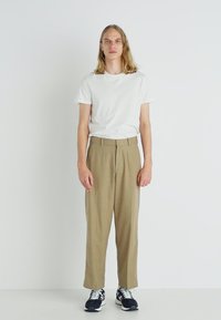 Beige, loose-fitting trousers with a high waist and front pleats, paired with a plain white t-shirt and black sports shoes.
