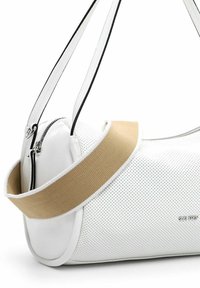 Weiße perforierte Lederhandtasche mit einem beigefarbenen Canvas-Trageriemen. Sie verfügt über einen Reißverschluss und hat eine abgerundete Form. Schwarze Akzente am Trageriemen.