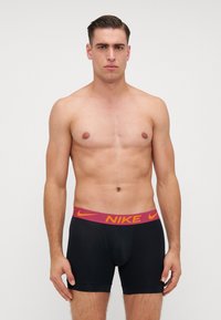 Melnas bokseru bikses ar kontrastējošu sarkanu un oranžu Nike logo jostu. Materiāls šķiet gluds, ar pieguļošu dizainu un īsu garumu.