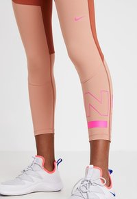 Legíny Nike v kombinaci béžové a hnědé barvy, s růžovým logem a těsným střihem, doplněné světle šedými sportovními botami.