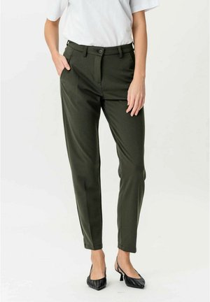 Nő sötétzöld, szabott nadrágban és fekete, hegyes orrú, pántos magassarkúban, egyik kezét a zsebében tartva, fehér felsőben áll.