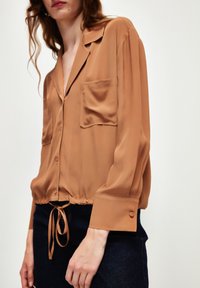 Braune Satin-Hemd mit Knöpfen, zwei Brusttaschen, einem Kordelzugsaum und langen Ärmeln. Weicher Stoff mit lockerer Passform.