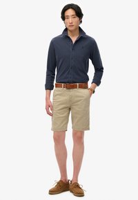 Marineblauwe shirt met lange mouwen, kaki shorts, bruine riem en bruine suède schoenen. Het shirt heeft een knoopsluiting en de shorts zijn knielang.