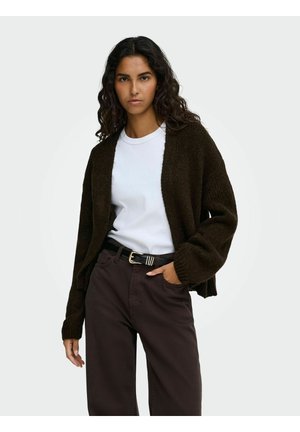 Femme aux longs cheveux bouclés portant un cardigan marron foncé ouvert, un t-shirt blanc, un pantalon marron foncé et une ceinture noire avec une boucle dorée.