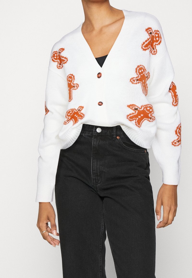 Cardigan blanc avec motifs d'homme en pain d'épices orange, boutonné devant, porté avec un jean noir taille haute sur une personne debout.