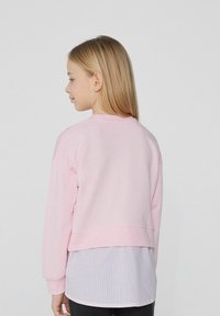 Helles rosa Sweatshirt mit einem cropped Design, gerippten Bündchen, weicher Textur, getragen über einem blau-weiß gestreiften Hemd und dunklen Hosen.