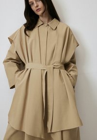Trench coat bej cu mâneci supradimensionate, guler, închidere frontală cu butoane și talie legată. Fabricat dintr-un material neted și ușor.