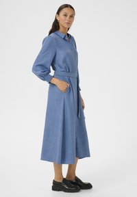 Robe bleu clair à manches longues, boutonnée, avec une taille ceinturée, des poches latérales et une texture douce. Accompagnée de chaussures noires à semelles épaisses.