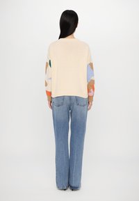 Kremowy sweter z kolorowymi abstrakcyjnymi wzorami na rękawach, zestawiony z niebieskimi, wysokim stanem jeansami. Miękka dzianina o luźnym kroju.