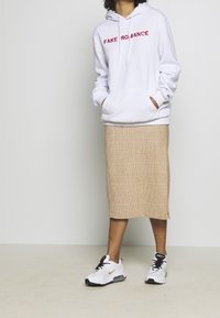 Vit hoodie med "FAKE ROMANCE" i röd brodering, ihopparad med en beige rutiga kjol och vita Nike sneakers. Avslappnad, lager-på-lager outfit.