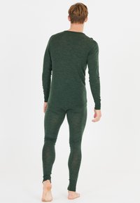 Man die blootsvoets staat, met zijn rug naar de camera, gekleed in een aansluitend donkergroen shirt met lange mouwen en bijpassende leggings voor een effen witte achtergrond.