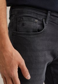 Crne traperice uskog kroja, s prednjim džepom s utisnutim logom, metalnim kopčanjem i suptilnom teksturiranom površinom.