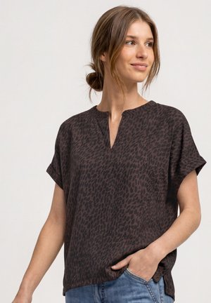 Femme aux cheveux châtain clair en chignon bas, portant un chemisier ample à manches courtes imprimé léopard brun foncé et un jean bleu, une main dans la poche.