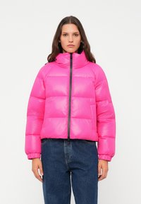 Veste doudoune rose avec col haut, fermeture éclair à l'avant et poignets élastiques. Présente un design matelassé et une coupe oversize, associée à un jean bleu.