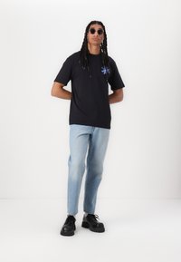 Sort t-shirt med kort ærme og blå blomstermotiv på brystet, parret med lyseblå jeans og sorte klodsede sko.