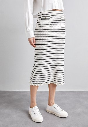 Vrouw draagt een witte midirok met zwarte horizontale strepen, voorzakken met knopen, een gerimpelde zoom, een wit overhemd en witte sneakers.