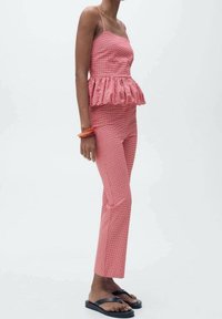 Rode gingham peplumtop met dunne straps, gecombineerd met bijpassende aansluitende broeken. Het model draagt zwarte sandalen en oranje armbanden.