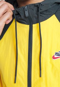 Yellow jacket with black hood. Features a central zip closure, drawstrings, and a small red and white logo detail on the chest.
