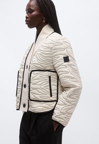 Sieviete ar pītām matu bizēm, valkājot krēmkrāsas izšūtu jaku ar melniem apšuvumiem un lielām kabatām, stāv profilā ar rokām melno biksu kabatās.