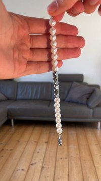 Pērļu rokassprādze ar apaļām baltām pērlēm, starp kurām ir metāliski pelēki akcenti. Vienā galā ir aizdare, attēlots rokā.