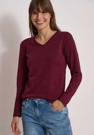 Femme souriante portant un haut rouge foncé à manches longues avec un col en V et un jean bleu, une main dans la poche, sur un fond uni.