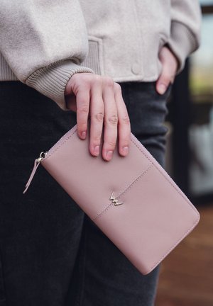 Pink leather wallet with a zip closure, featuring a gold accent and clean stitching. Held in a hand, worn with a grey sweater and dark jeans.