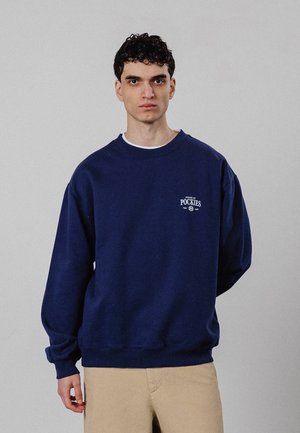 Jonge man met krullend donker haar die een marineblauwe trui draagt met de tekst "House of Pockies" en beige broek tegen een effen achtergrond.