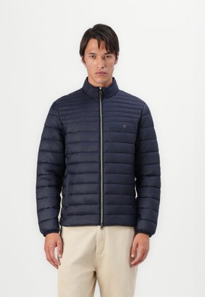 Navy puffer jacket with a high collar, horizontal quilted design, full front zipper, and elastic cuffs. Worn over beige pants.
