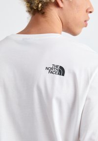 Weißes Baumwoll-T-Shirt mit einem schwarzen "The North Face"-Logo im oberen Rückenbereich, das eine glatte Textur und ein lässiges Design zeigt.