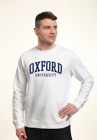 Vit sweatshirt med "OXFORD UNIVERSITY" i marinblå bokstäver, rund hals, långa ärmar och en mjuk bomullstextur.