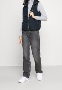 Granatowa pikowana kamizelka z zamkiem z przodu, z fakturą skośnych przeszyć, w zestawieniu z ciemnoszarymi dżinsami i białymi sportowymi butami. Dopasowany, bezrękawnik.