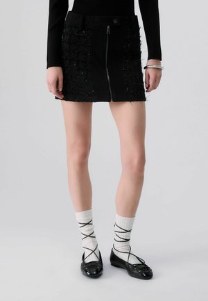 Femme portant une mini-jupe noire texturée avec fermeture éclair à l'avant, des chaussettes blanches avec lacets noirs croisés et des ballerines noires.